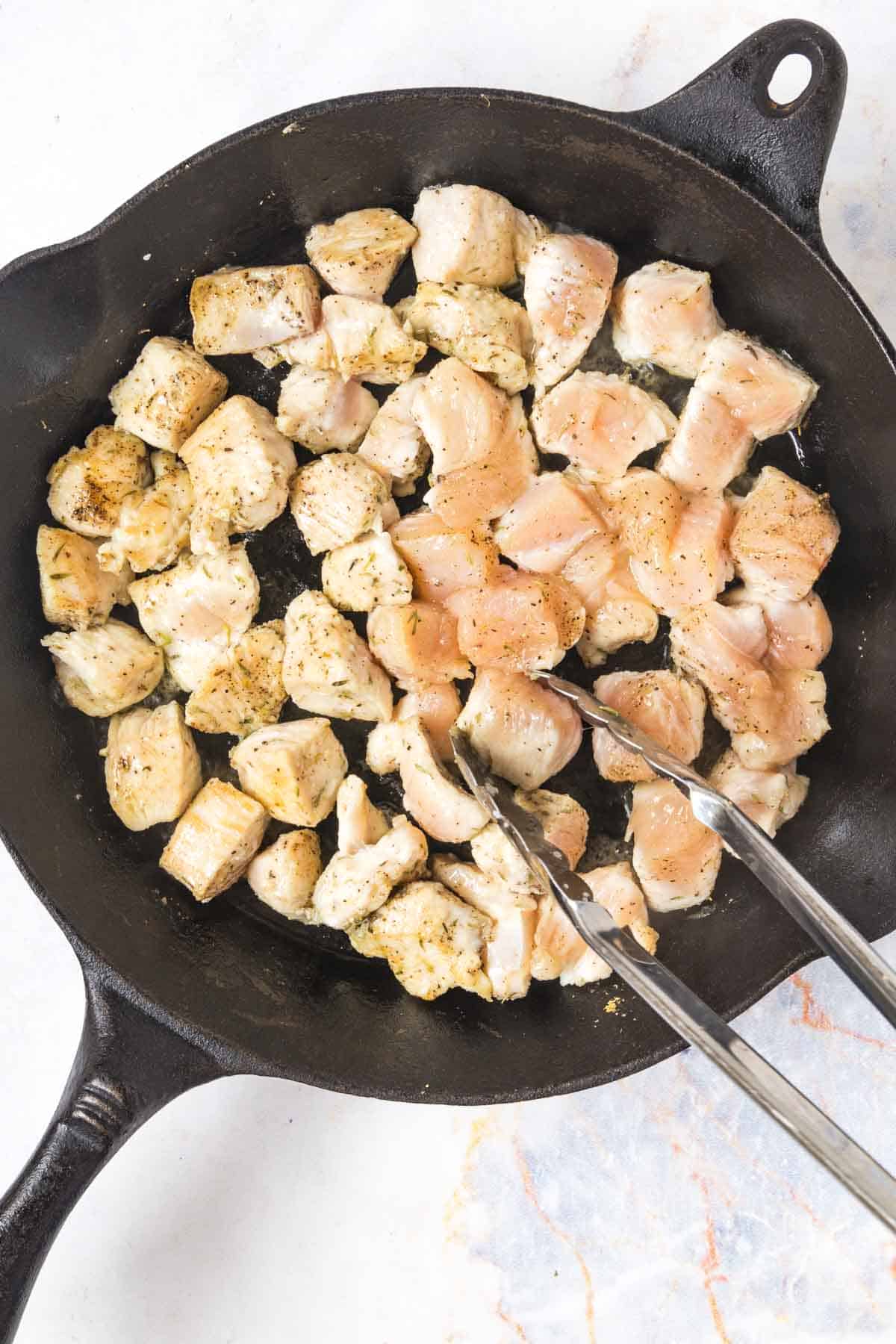 tongs flipping chicken bites in a cast iron skillet