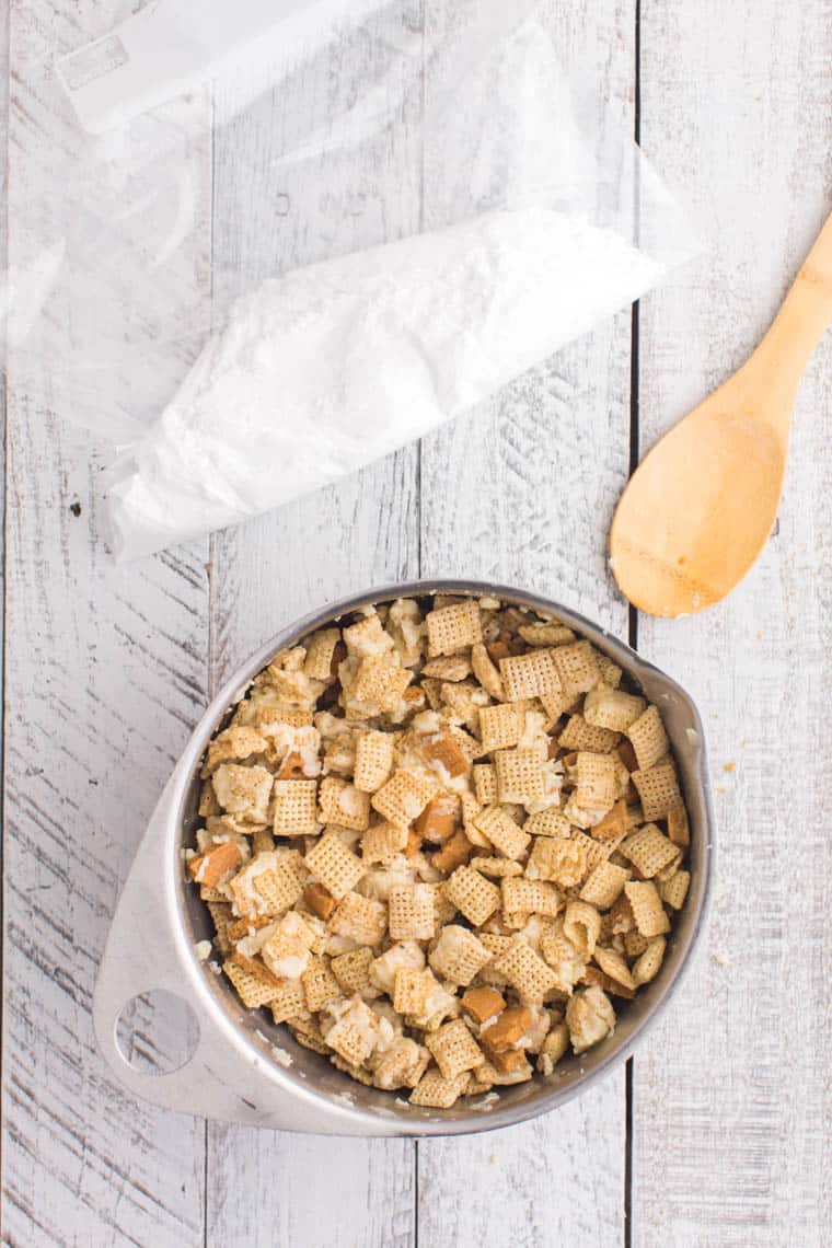 chex cereal and broken graham crackers coated in white chocolate in a bowl with a bag filled with powdered sugar