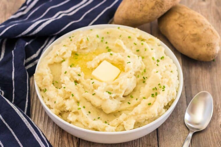 mashed potatoes in a bowl topped with a partially melted pat of butter and minced chives on a blue dish towel with two russet potatoes next to it