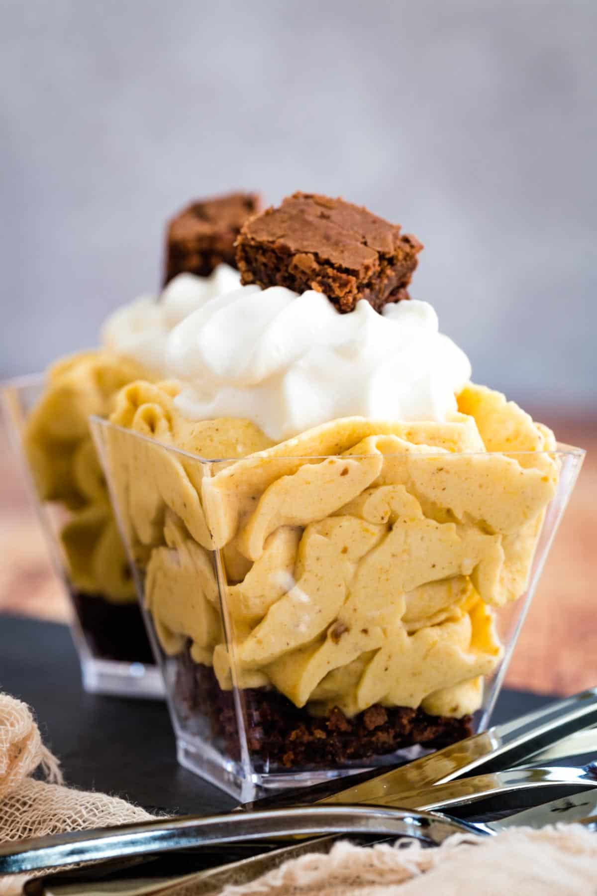 a mini pumpkin cheesecake mousse parfait topped with whipped cream and a small piece of brownie with another one behind it on a slate platter with mini spoons