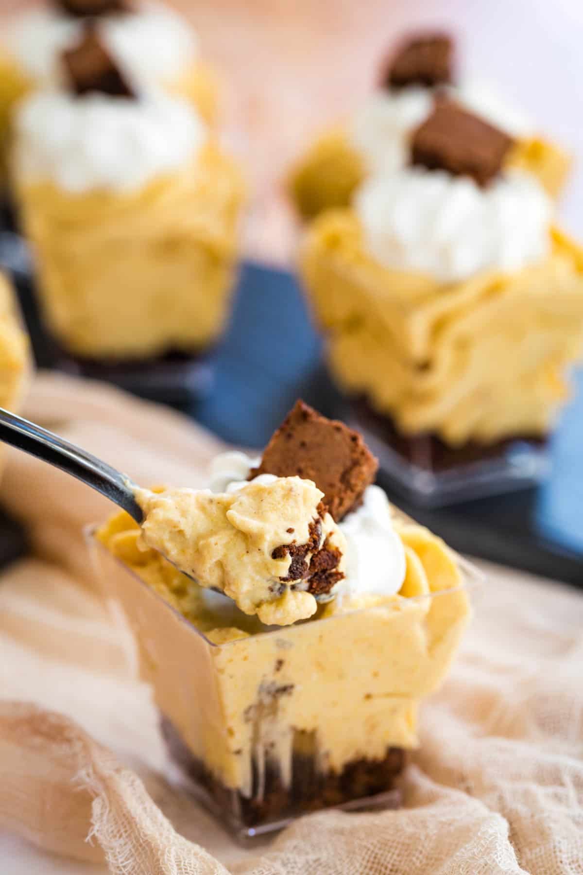 bite of pumpkin cheesecake mousse and brownie on a mini spoon with a bunch of parfaits in the background
