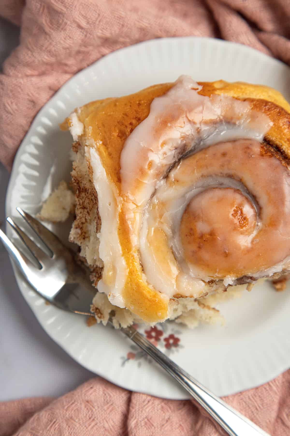 Looking down at a glazed gluten free cinnamon roll on a plate with a fork on top of a pink cloth napkin.