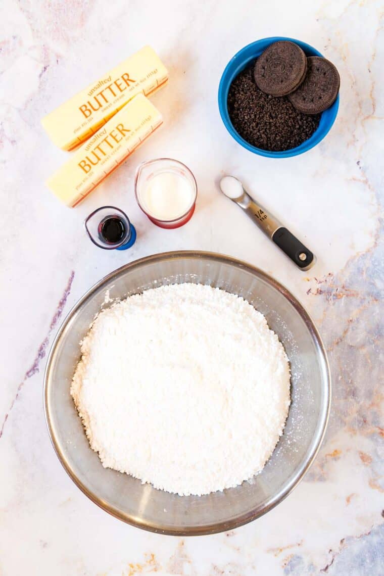 metal mixing bowl with powdered sugar, two sticks of butter, a one-quarter teaspoon of salt, two small beakers of vanilla and heavy cream, and a bowl of cookie crumbs with two gluten free oreos on top