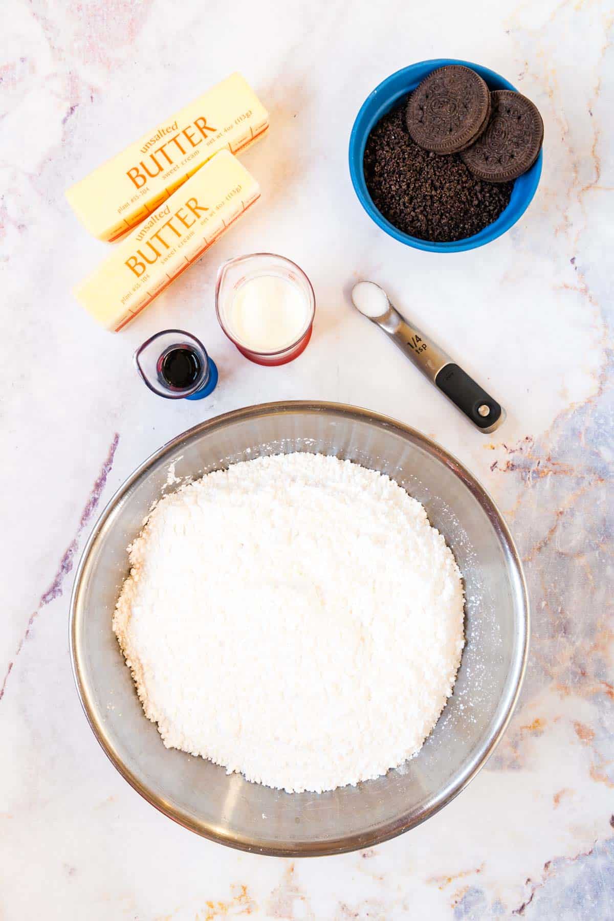 A metal mixing bowl with powdered sugar, two sticks of butter, a one-quarter teaspoon of salt, two small beakers of vanilla and heavy cream, and a bowl of cookie crumbs with two gluten free oreos on top.