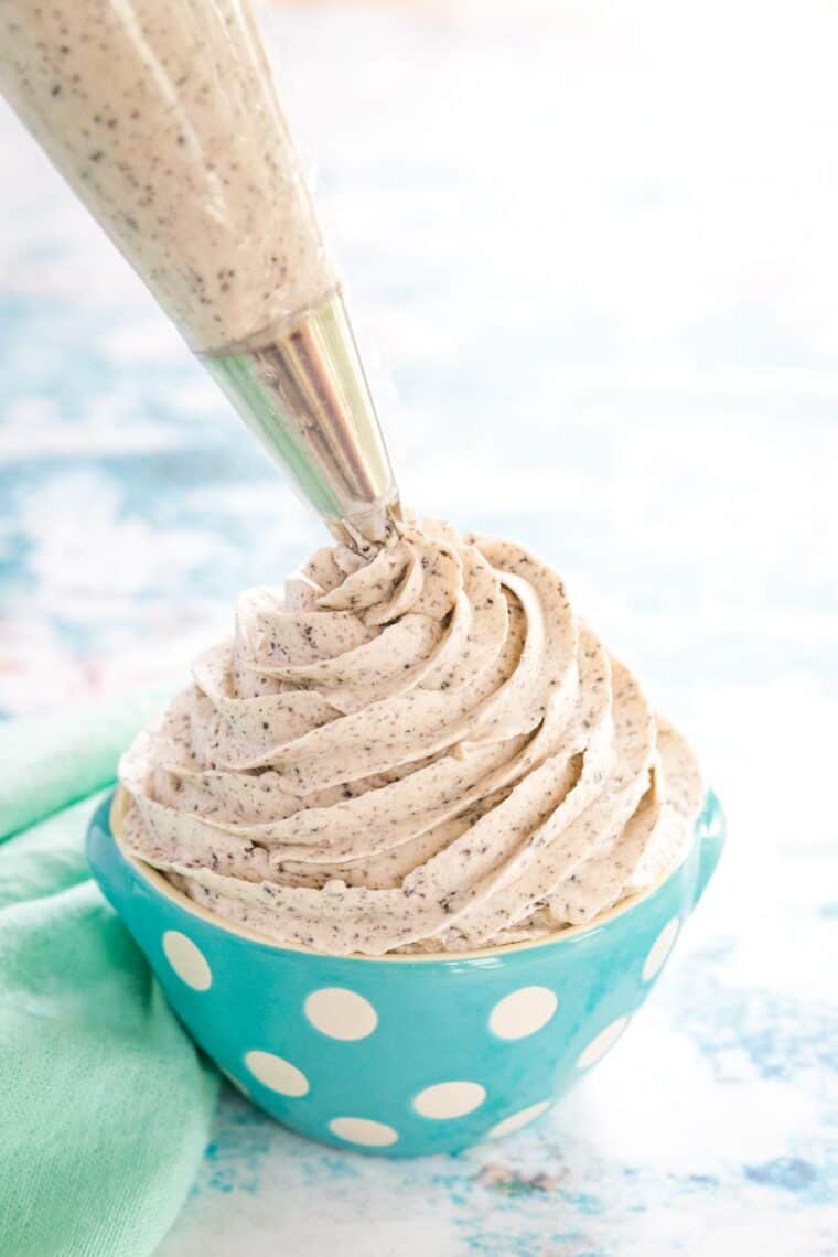 a piping bag with a star tip piping a swirl of oreo frosting into a blue bowl with white polka dots next to a mint green napkin