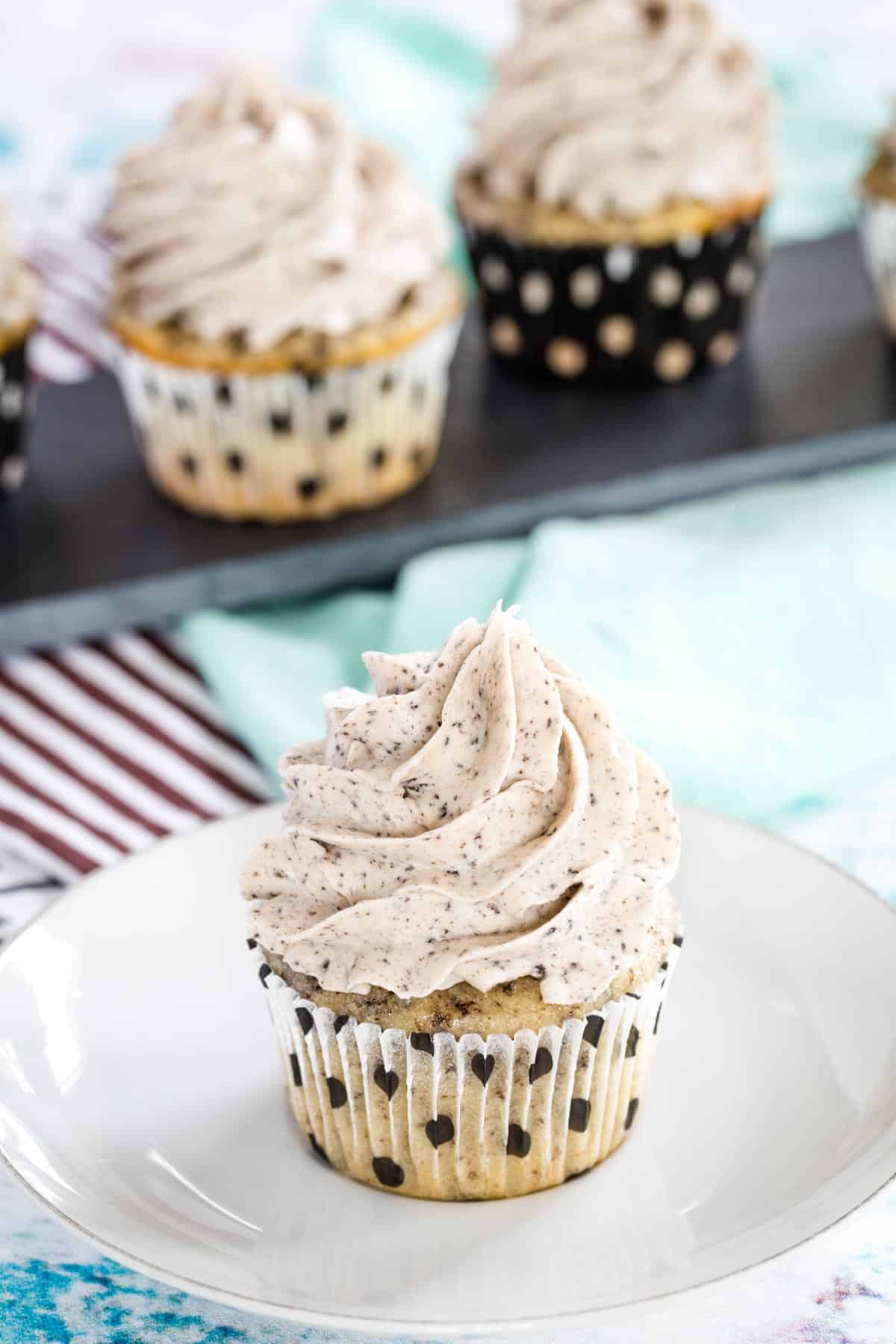 an oreo cupcake with a white cupcake liner with black polka dots on a white plate with three more cupcakes on a platter behind it