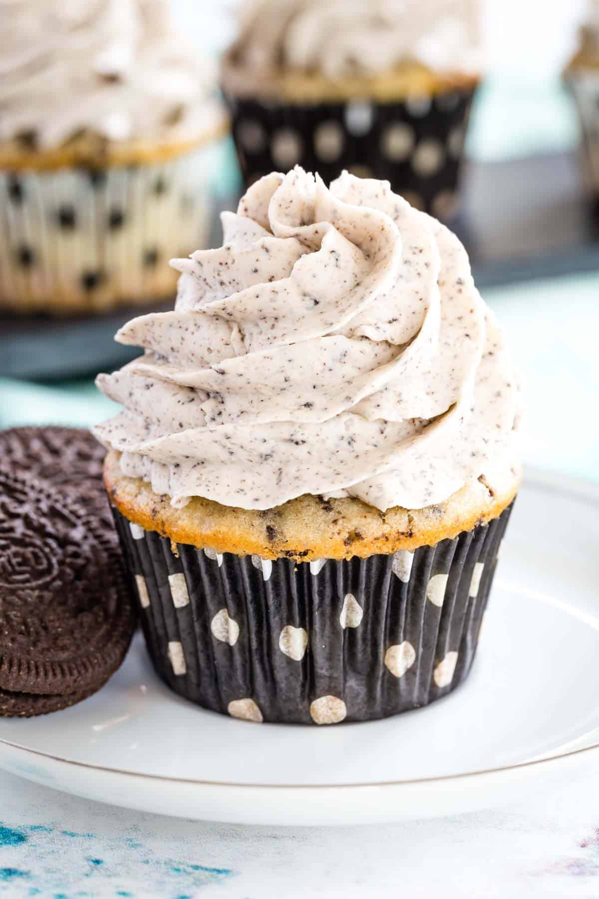 cookies and cream cupcake with a swirl of frosting flecked with cookie pieces on a white plate with two gluten free oreos