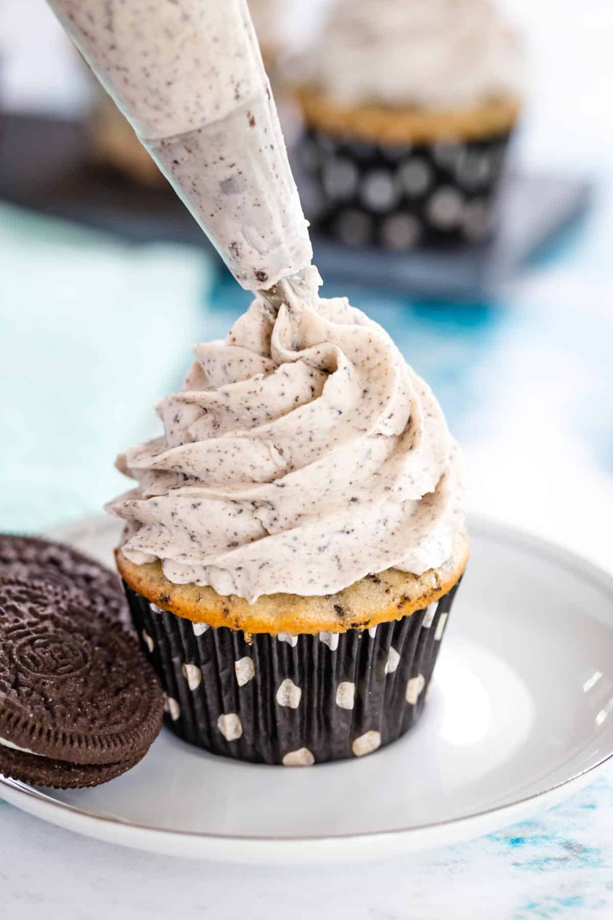 cookies and cream frosting bring piped out of a pastry bag into a swirl on an oreo cupcake sitting on a plate next to two gluten free oreos