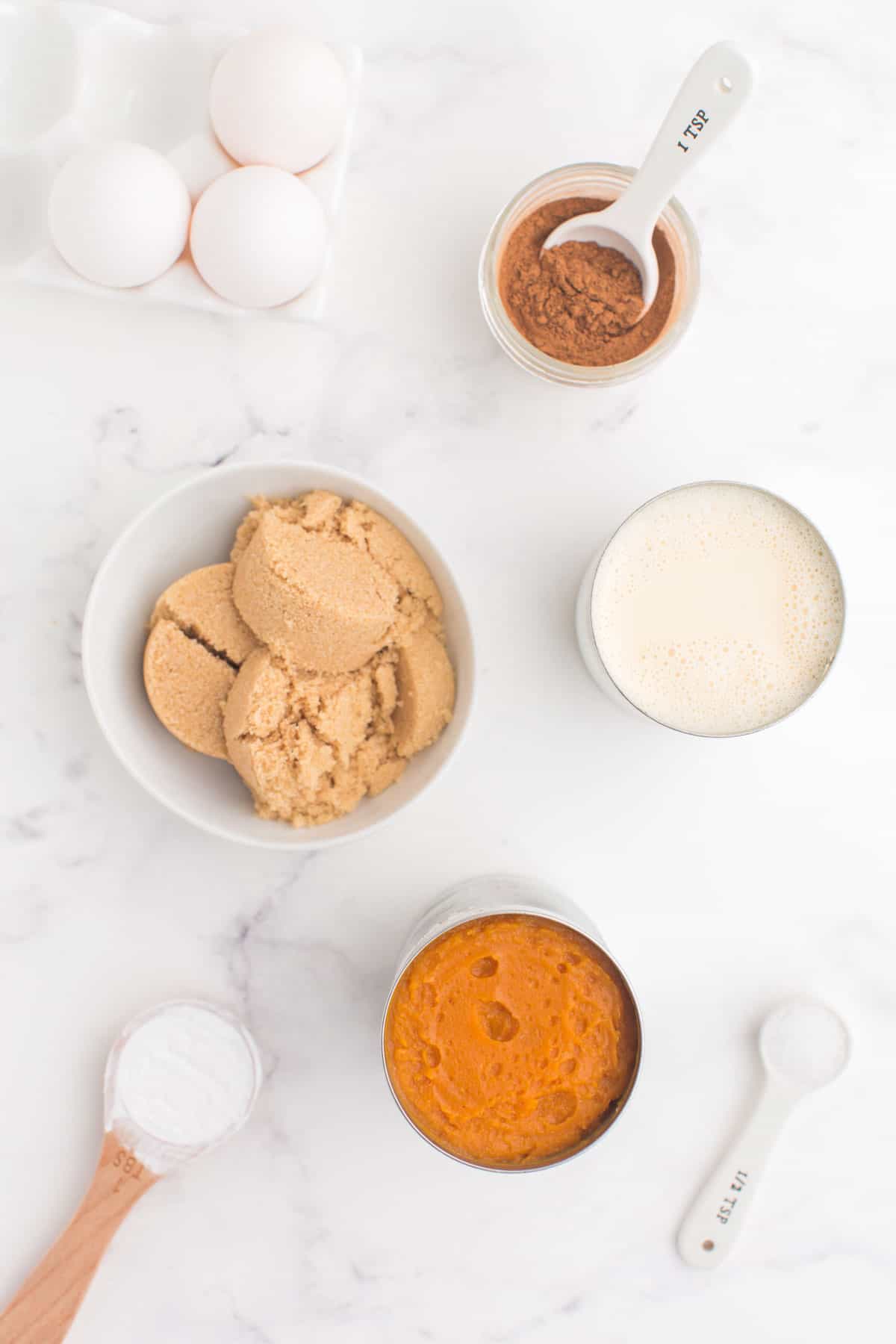 a can of pumpkin puree, a can of evaporated milk, three eggs, a bowl of brown sugar, and measuring spoons with cornstarch , salt, and pumpkin pie spice