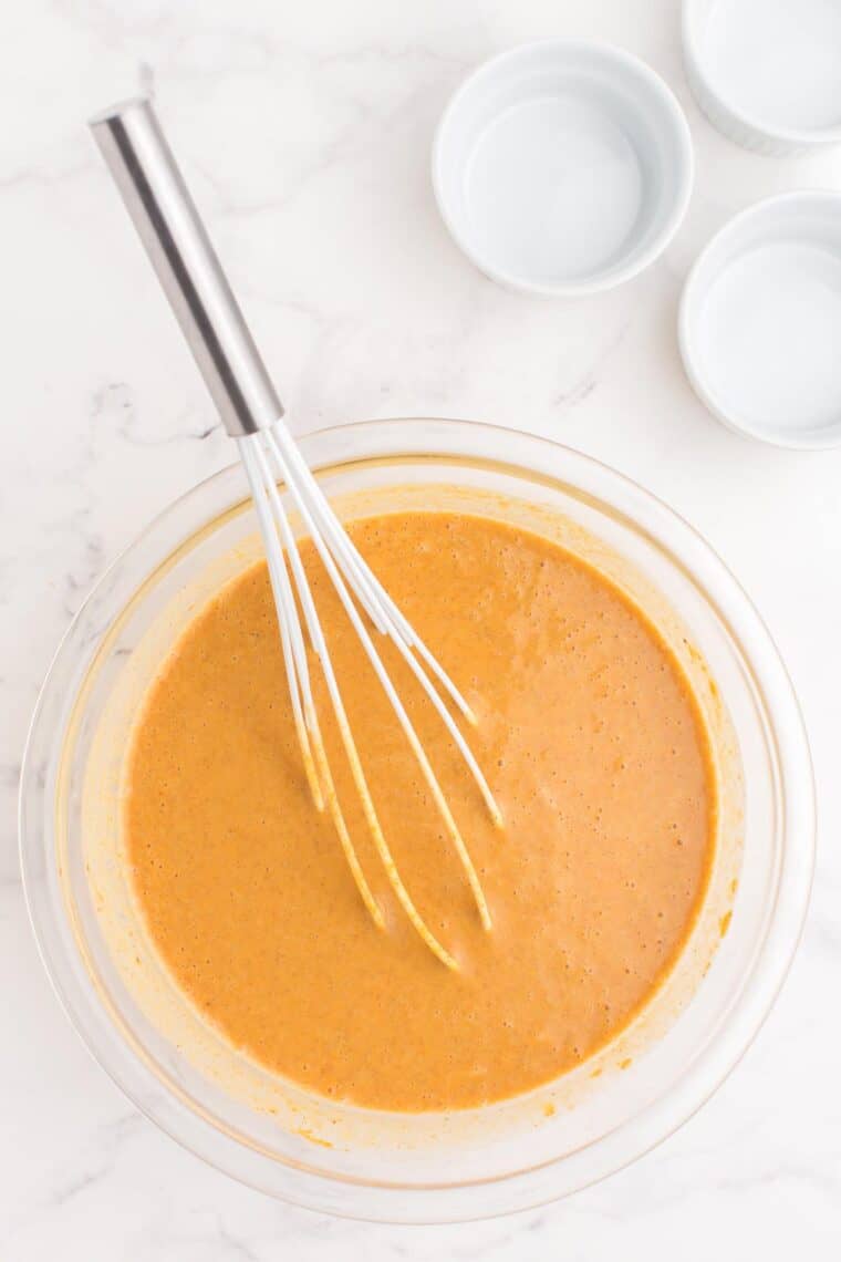 uncooked pumpkin pie filling mixture in a bowl with a whisk next to some ramekins