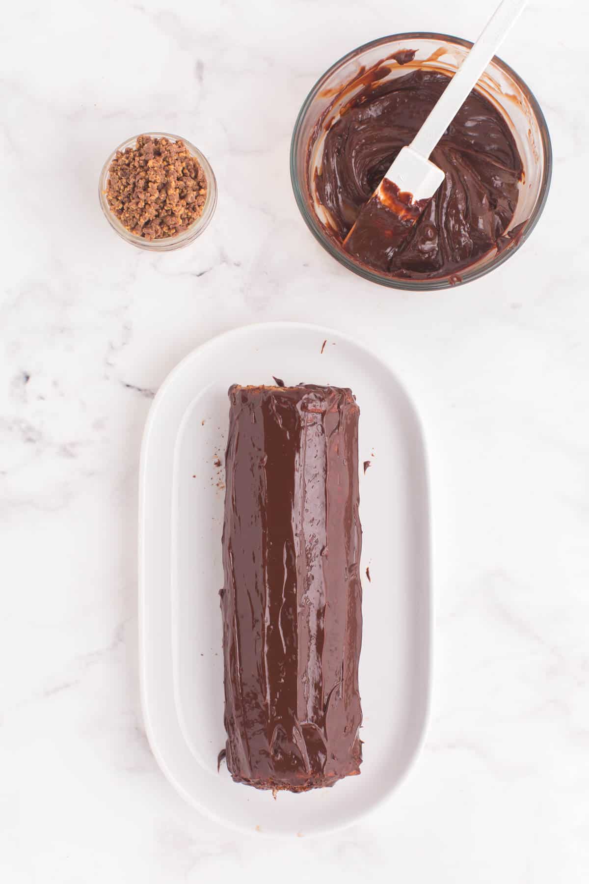 chocolate cake roll with chocolate ganache spread on the outside plus a bowl of chopped peanut butter cups and a bowl with the rest of the ganache