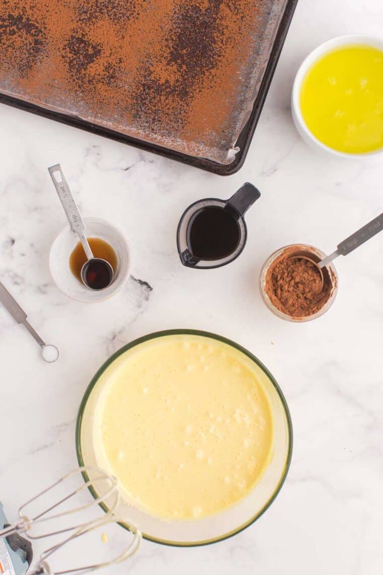 bowl of egg yolks and sugar that have been beaten until pale yellow on a table with bowls of vanilla, cooled coffee, egg whites, and cocoa powder