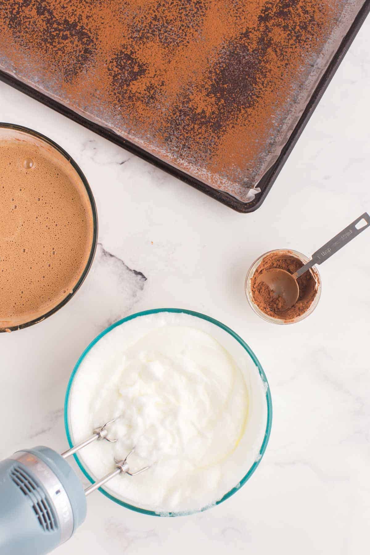 beater in a bowl of egg whites whipped until to stiff peaks plus the chocolate cake batter base in a bowl and a bowl of cocoa powder