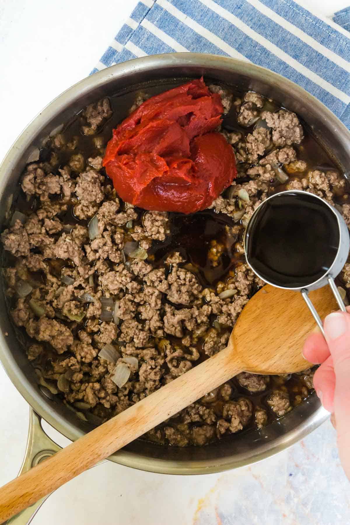 pouring worcestershire sauce into a skillet with cooked ground beef, beef broth, and tomato paste