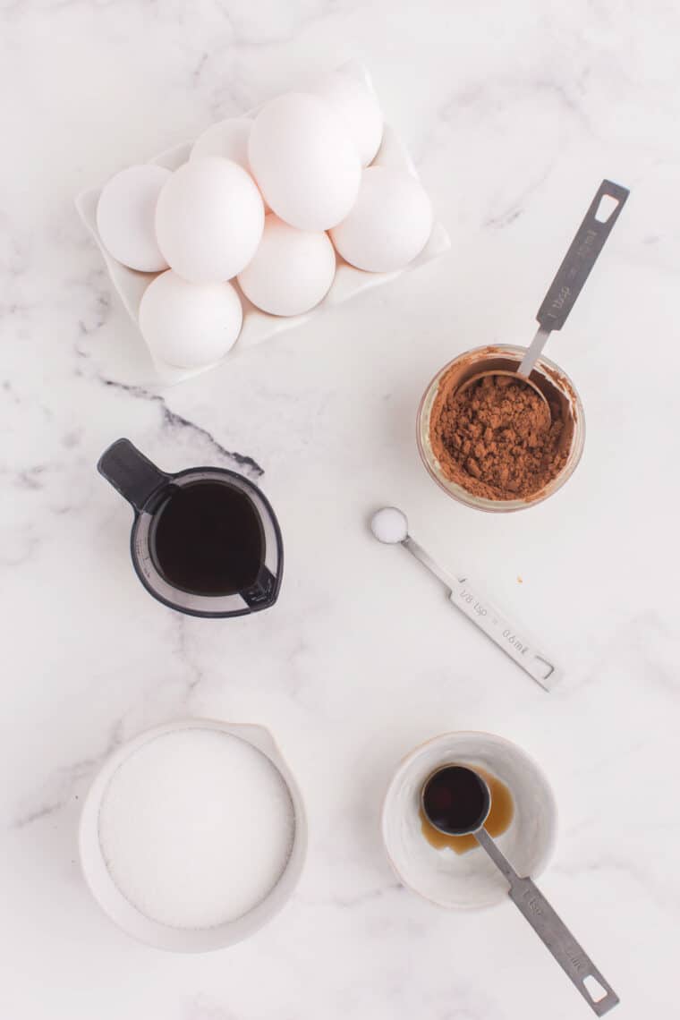 eight eggs, bowls of sugar, vanilla extract, and cocoa powder, a measuring spoon of salt and a measure cup with cooled coffee