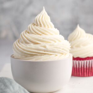 A bowl of cream cheese frosting with some piped on a cupcake behind it.