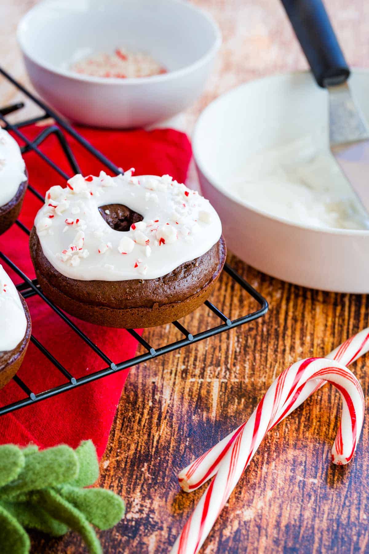frosted chocolate donut on the edge of a cooling rack next to bowls of icing and candy cane pieces