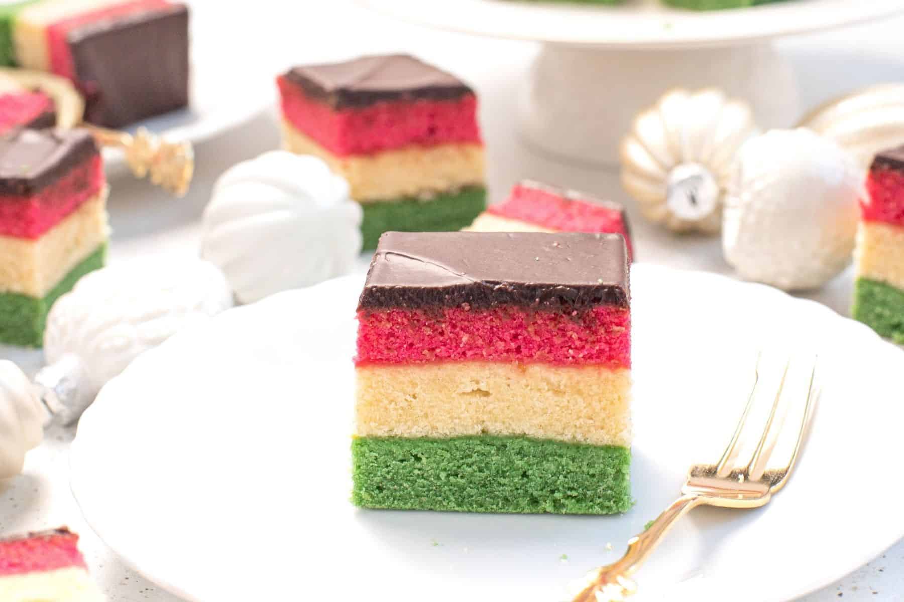 small square of a rainbow cookie on a white plate with a dessert fork