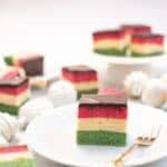 one piece of gluten free rainbow cookies on a plate with more on a cake stand