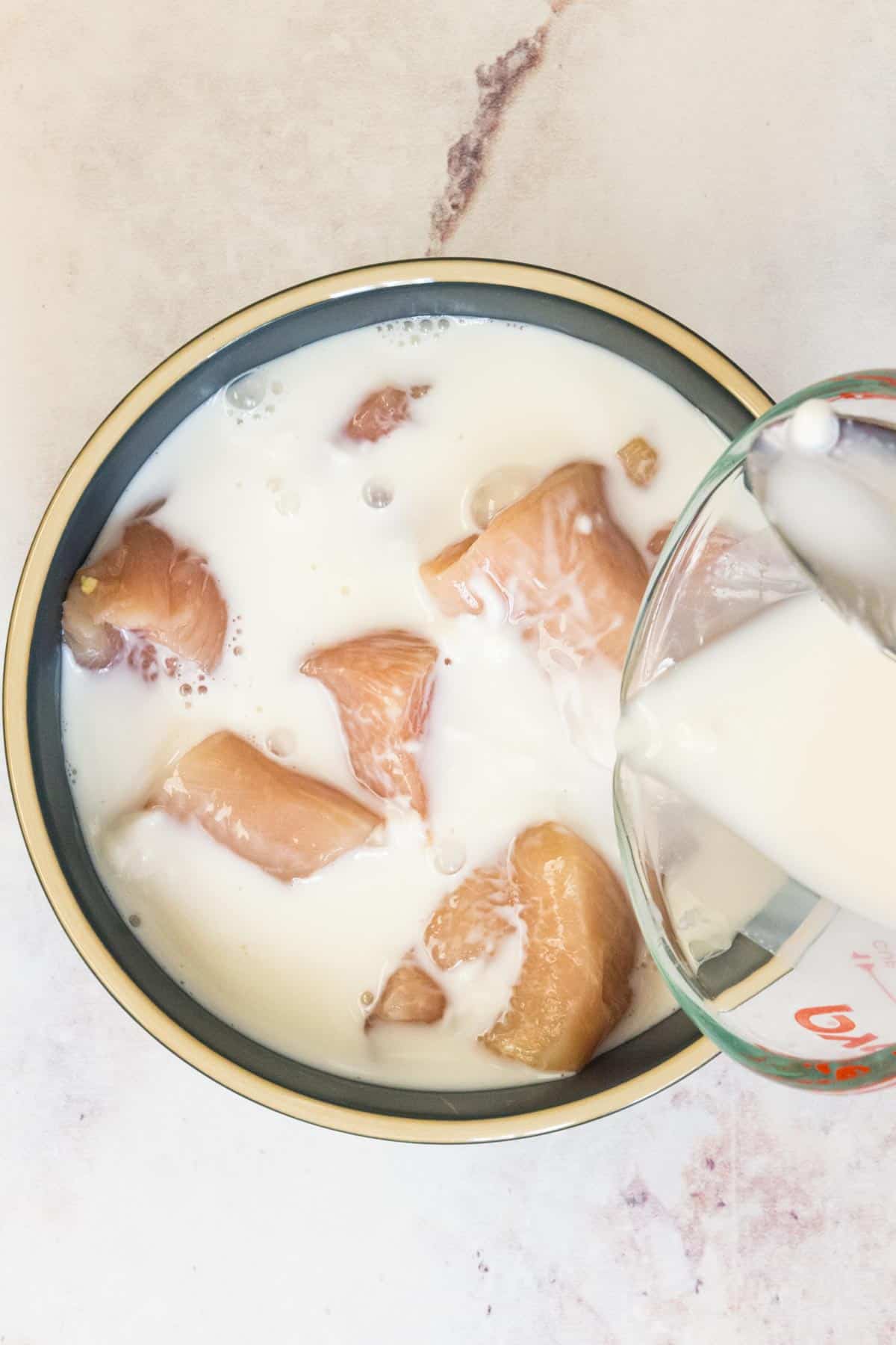 A marinade of plain Greek yogurt and milk is poured over a bowl of chicken pieces.