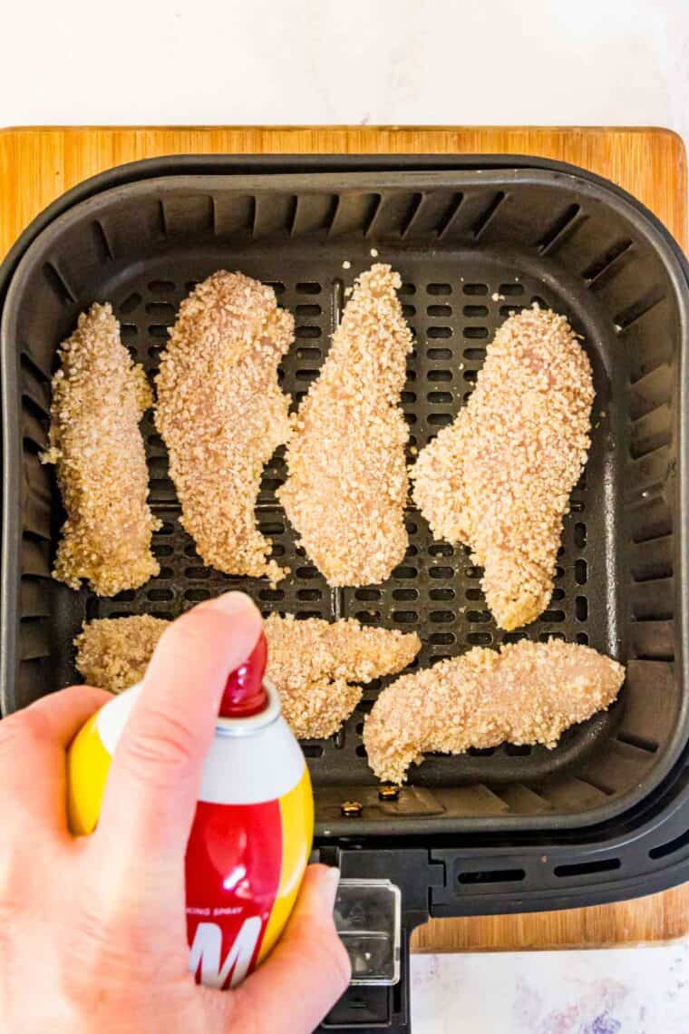 Chicken tenders in an air fryer are sprayed with cooking oil.