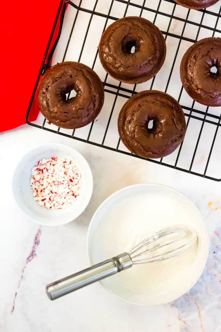 chocolate donuts on a rack, a bowl of white icing, and a bowl of chopped candy canes