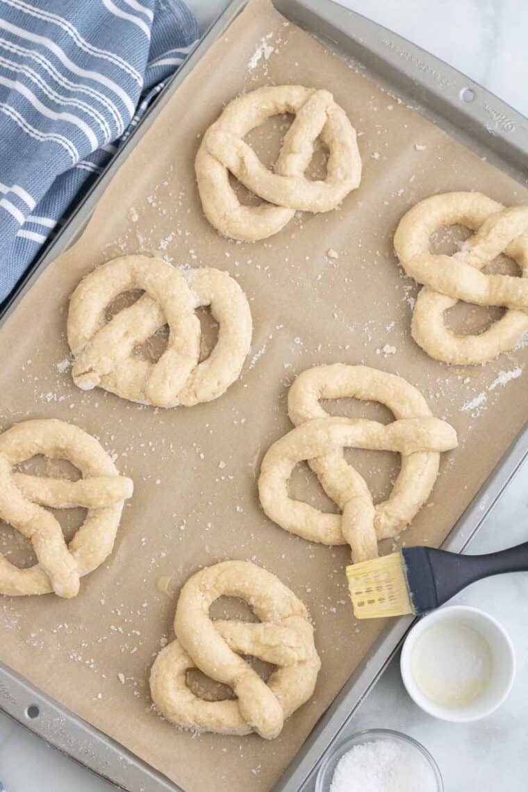 Twisted pretzel shapes on a baking sheet are brushed with melted butter.