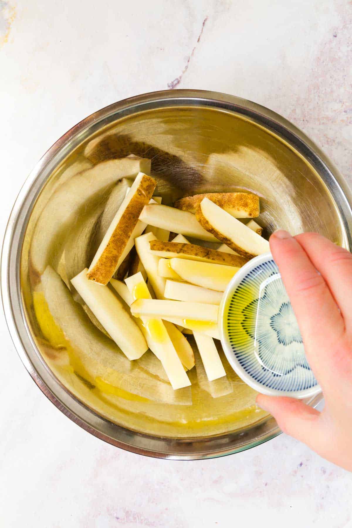 A tablespoon of oil being poured into a large bowl filled with unbaked french fries