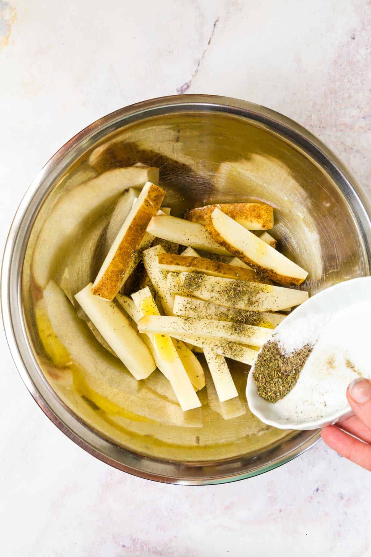 Salt, pepper and Italian seasoning being poured into a metal bowl with unbaked fries and oil