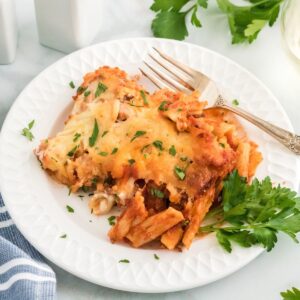 A scoop of baked ziti on a white plate with a fork and a sprig of parsley as a garnish.