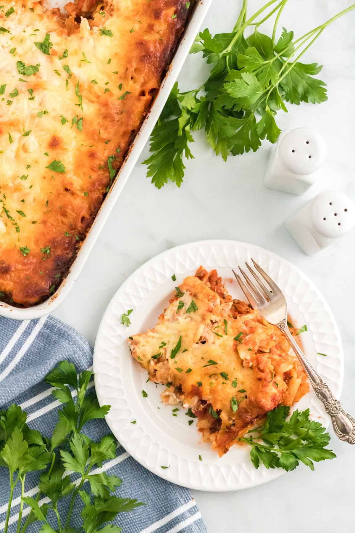 A slice of baked ziti on a plate next to a casserole dish.