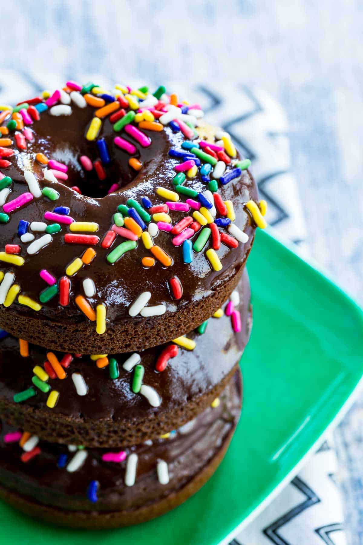 A stack of gluten-free chocolate donuts with rainbow sprinkles on a plate.