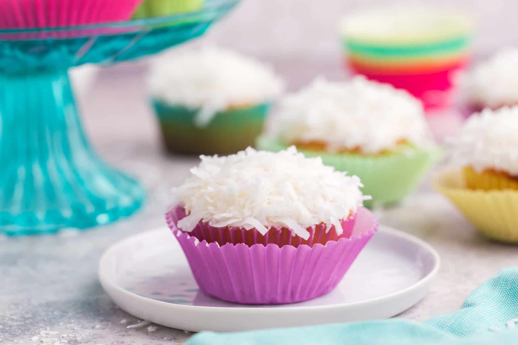 a coconut cupcake in a pink wrapper on a small plate with more cupcakes in the background.
