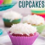 A cupcake with white frosting and shredded coconut on top in a pink wrapper on a small plate with more cupcakes in the background.
