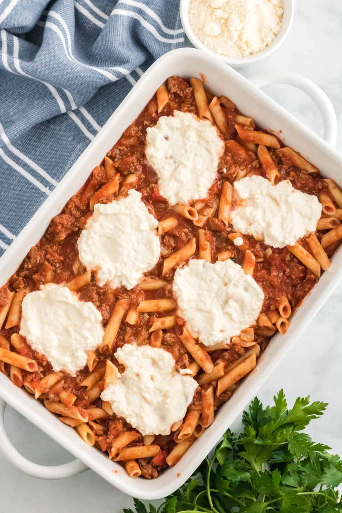 A layer of ziti pasta coated with meat sauce in a baking dish, topped with dollops of ricotta cheese.