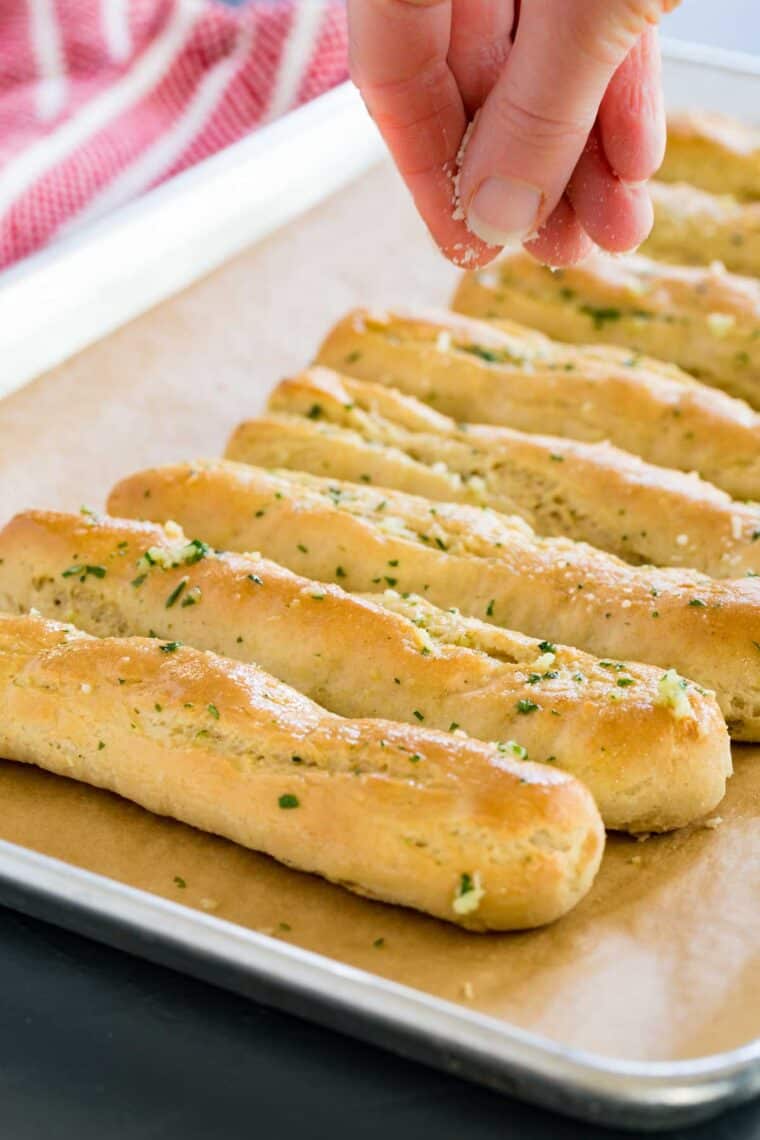 Sprinkling parmesan cheese over garlic breadsticks on a sheet pan.