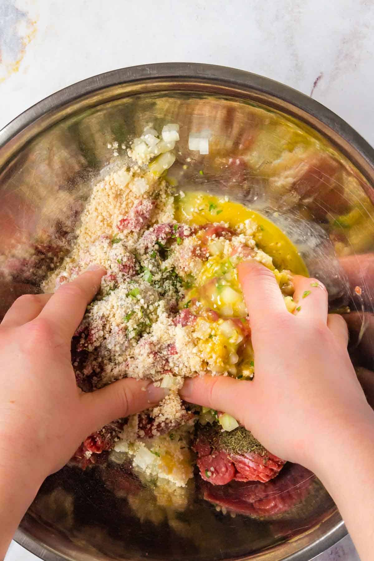 The meat mixture ingredients in a metal mixing bowl being combined by a pair of hands