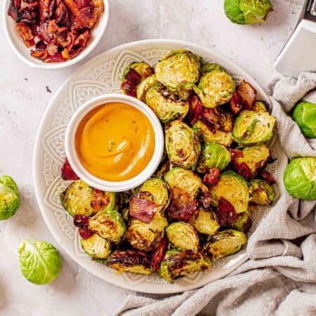 Brussels sprouts with bacon on a serving plate with dip next to a bowl of crispy bacon pieces
