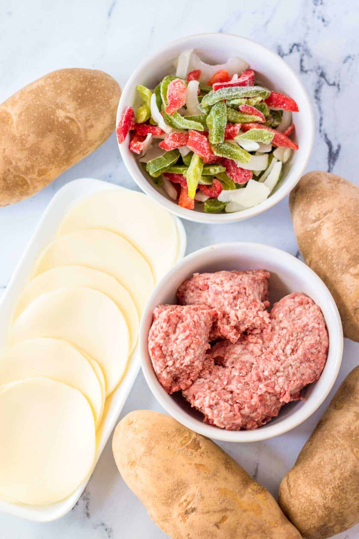 Bowls of ground beef and frozen peppers and onions, sliced of cheese, and four russet potatoes.