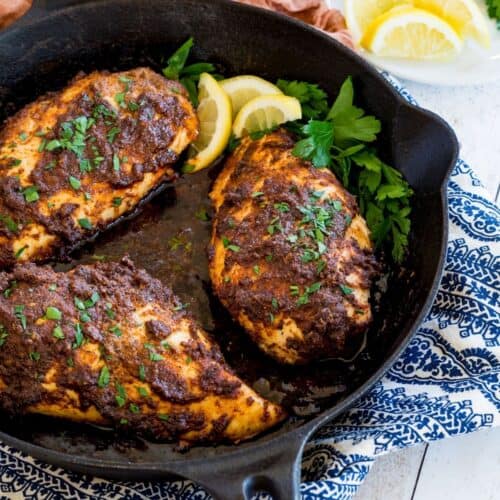 A cast iron skillet with three blackened chicken breasts garnished with parsley and lemon slices.