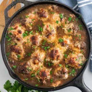 Meatballs in a brothy sauce with melted cheese in a cast iron skillet.