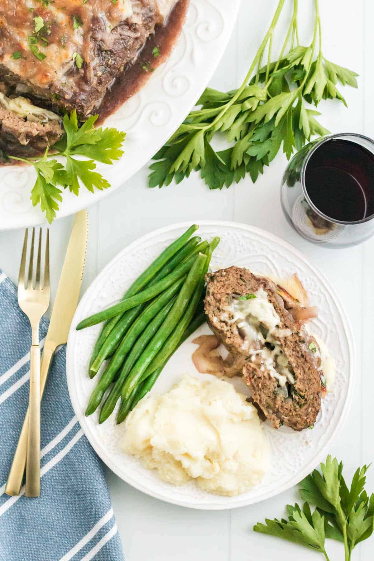 A serving of French onion stuffed meatloaf on a plate with veggies and potatoes beside a knife and fork