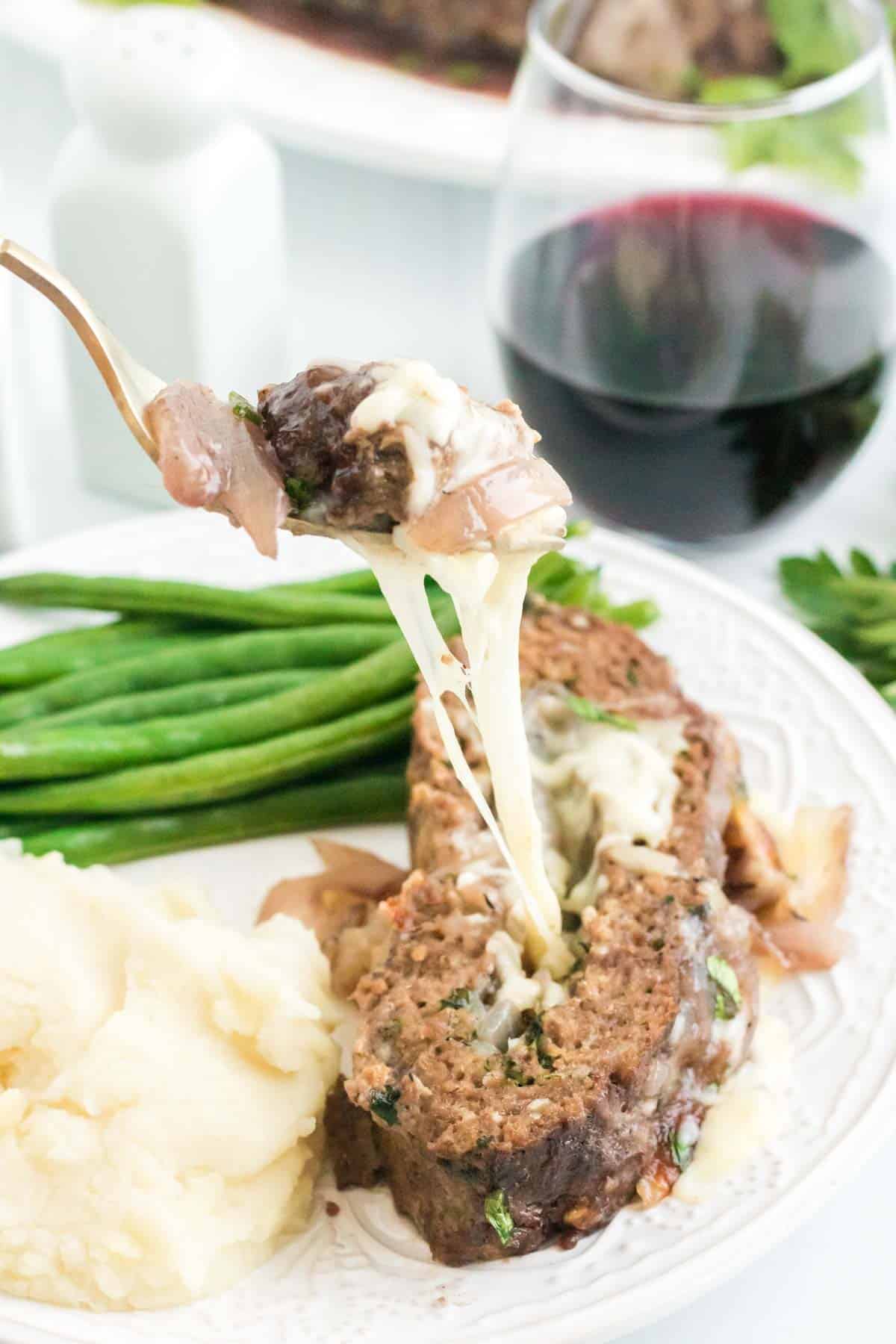 A fork holding a bite of meatloaf over a plate holding a slice of meatloaf, mashed potatoes and green beans