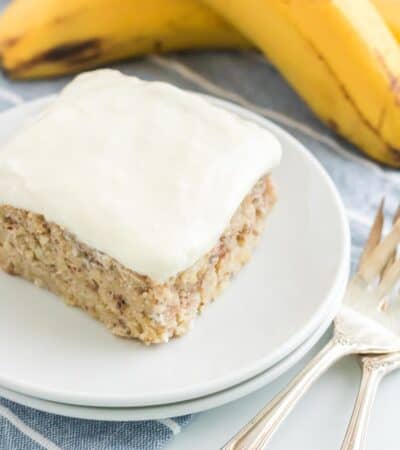 A piece of frosted cake on a plate with two bananas in the background