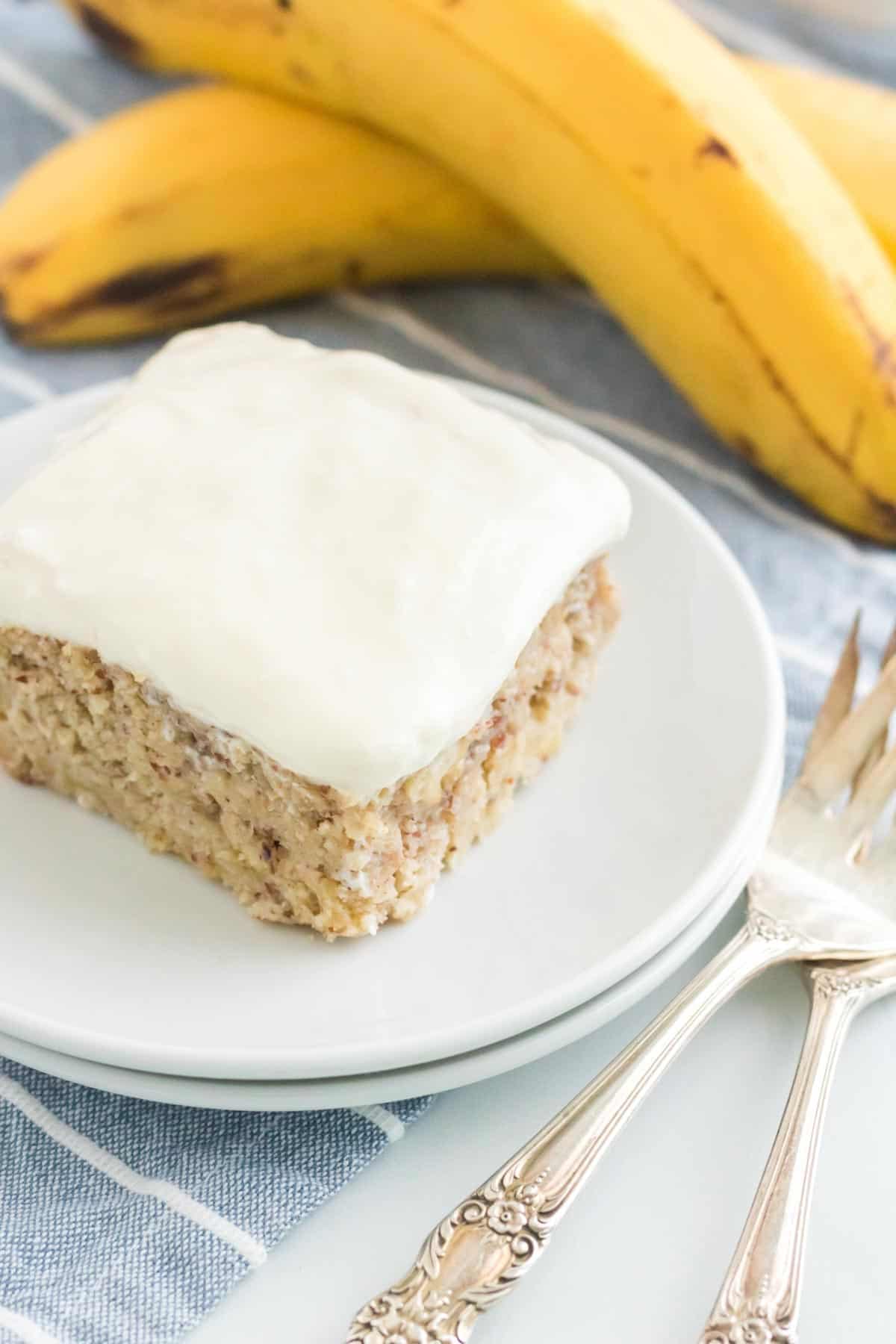 A piece of frosted cake on a plate with two bananas in the background