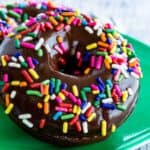 One gluten free chocolate donut with chocolate glaze and sprinkles leaning against another one ona green plate.