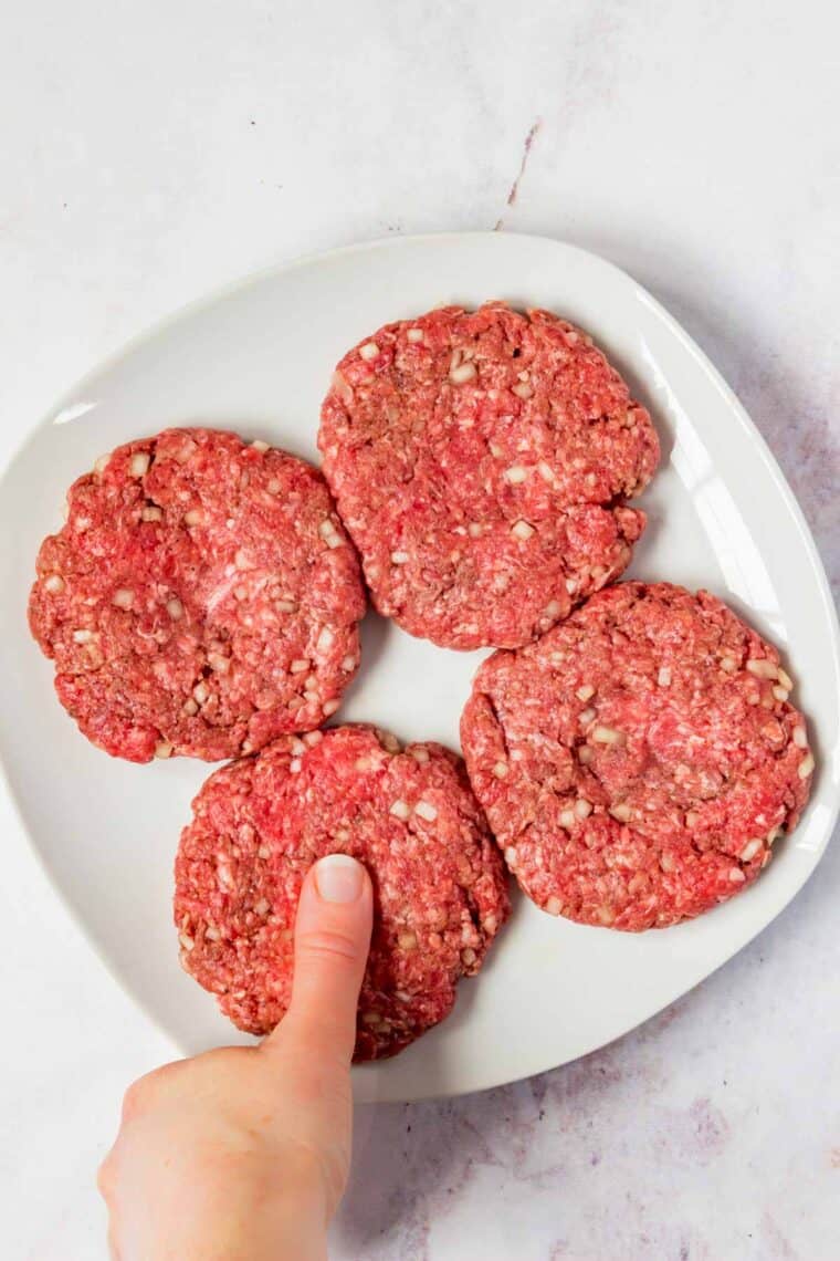 Four raw burger patties on a plate, making an indentation on one with a thumb.