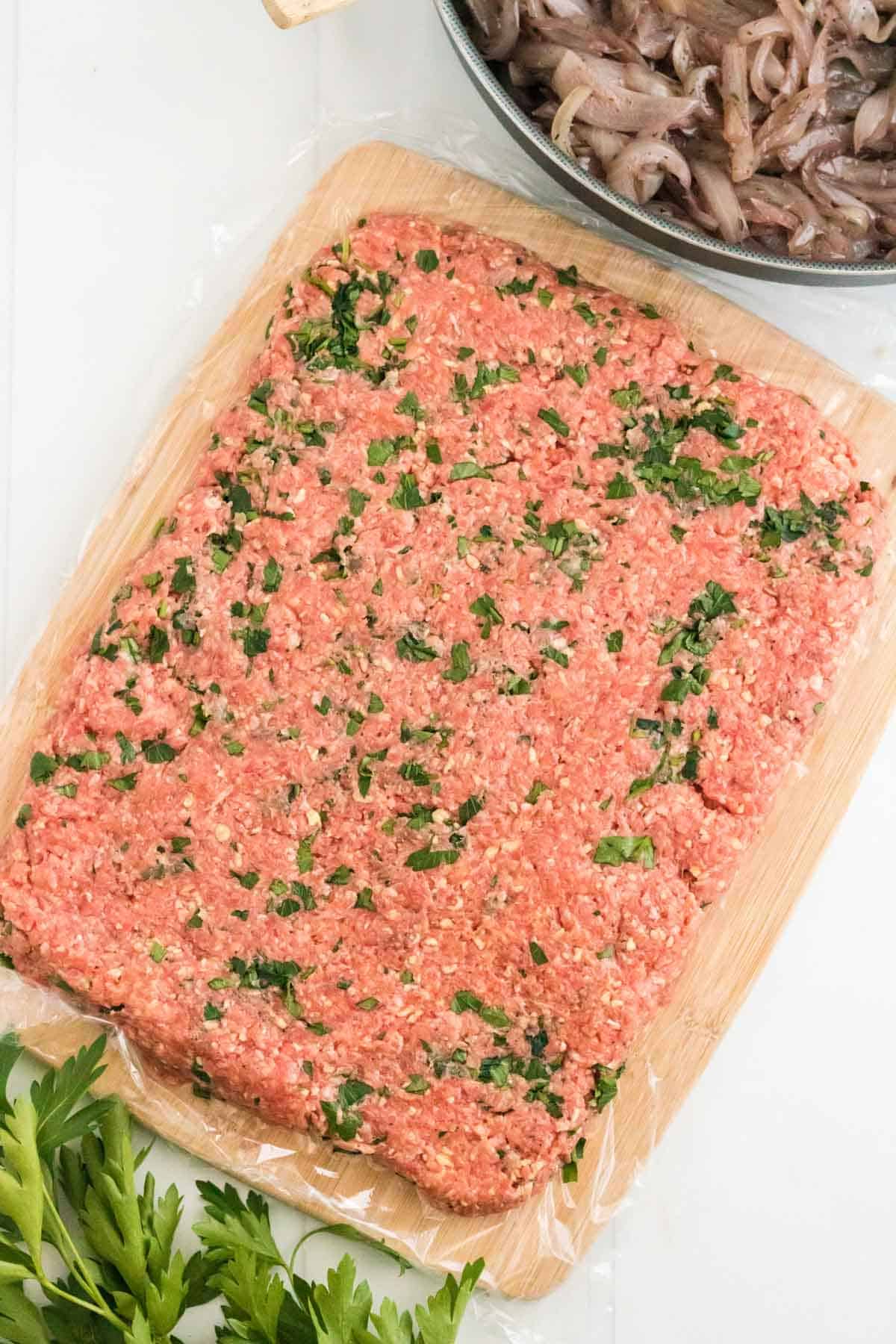 The meatloaf mixture in the shape of a rectangle on top of a cutting board lined with plastic wrap