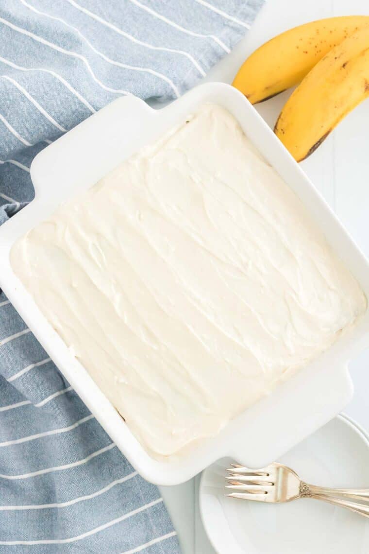 A frosted banana snack cake inside of a cake pan with a stack of plates on the counter next to it