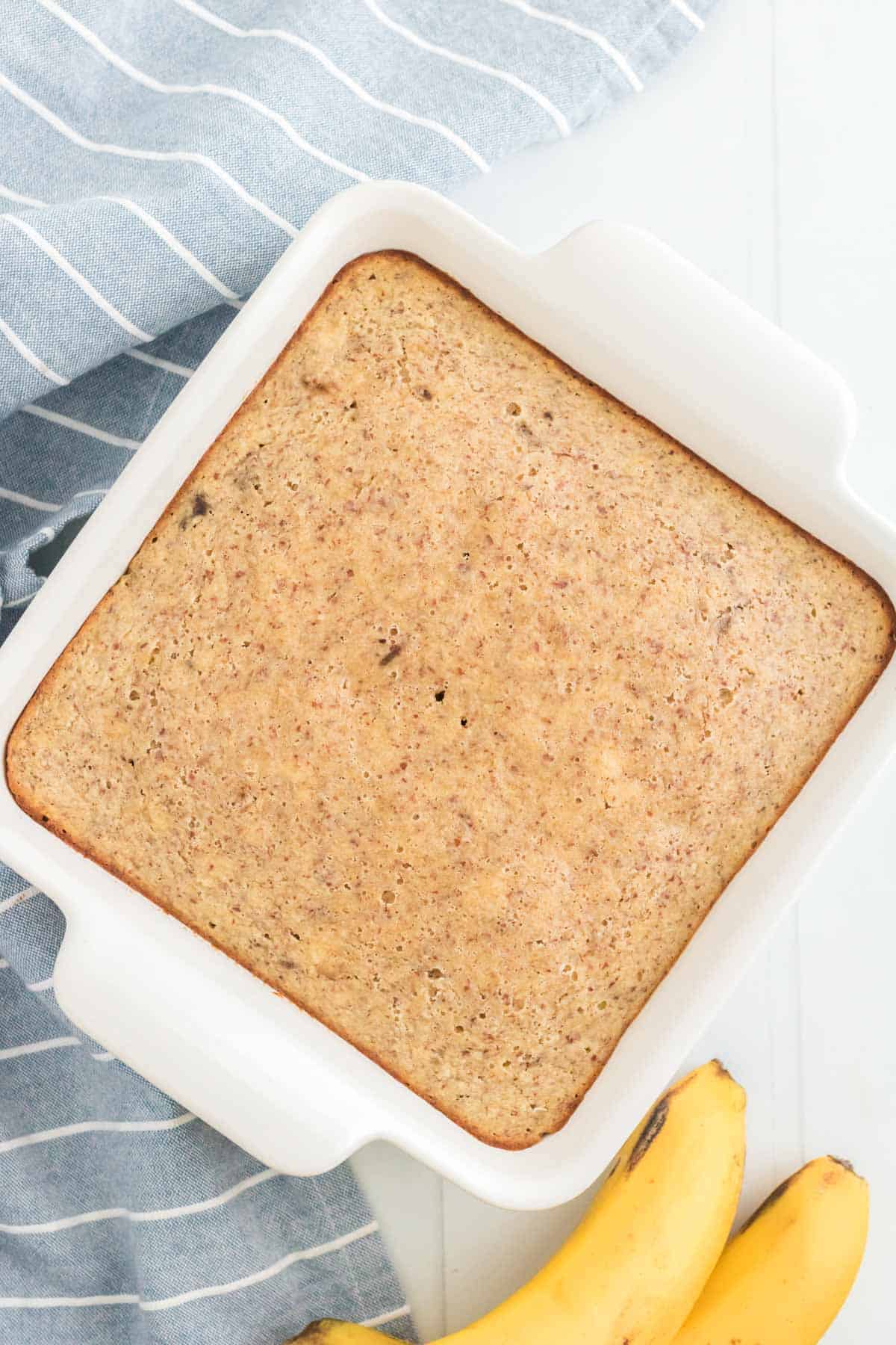 A baked banana snack cake inside of a square pan with two fresh bananas beside it